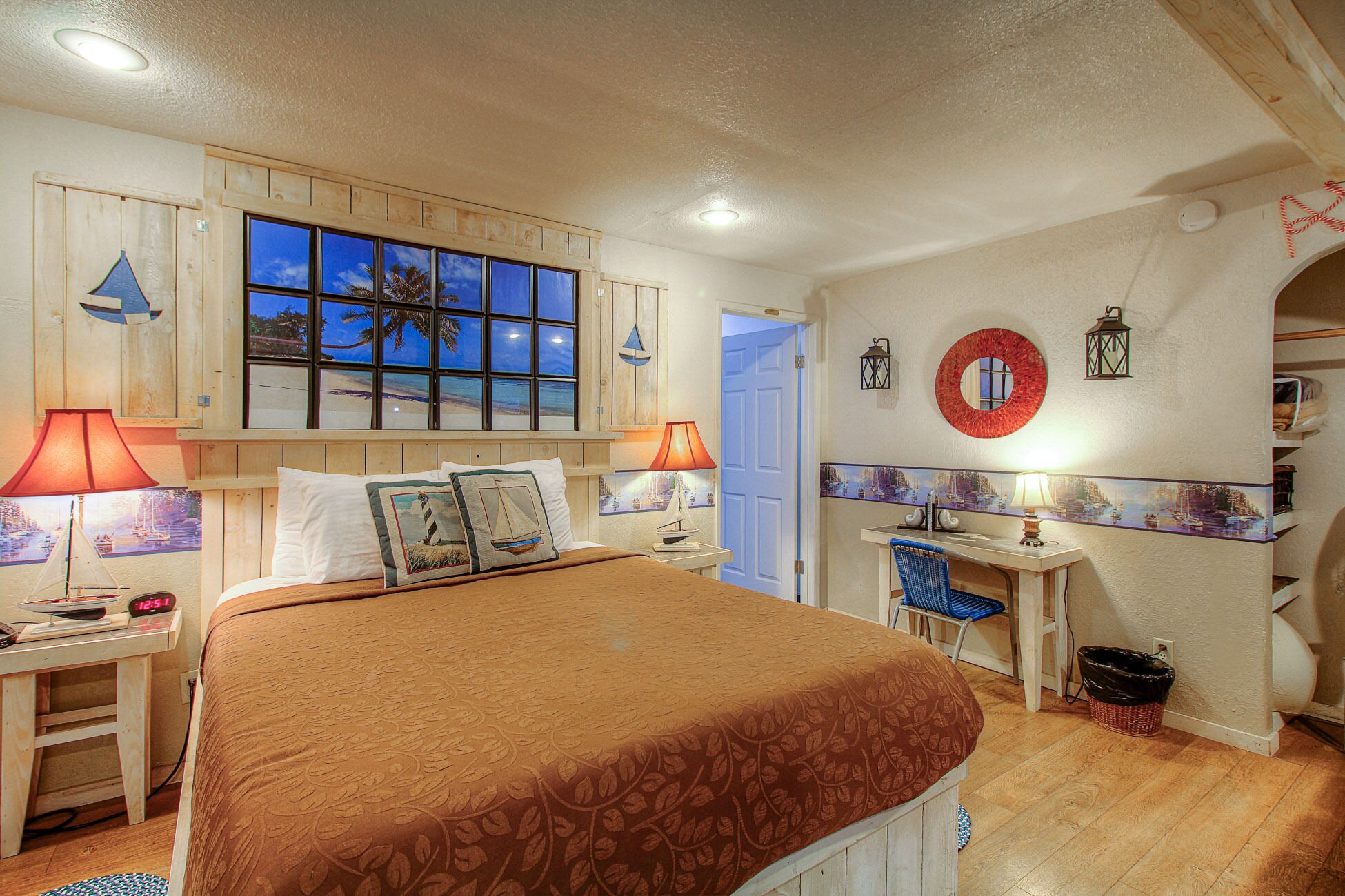 A bedroom with a large bed, two bedside lamps, a nightstand, a small desk, a chair, a wall-mounted mirror, and a door.