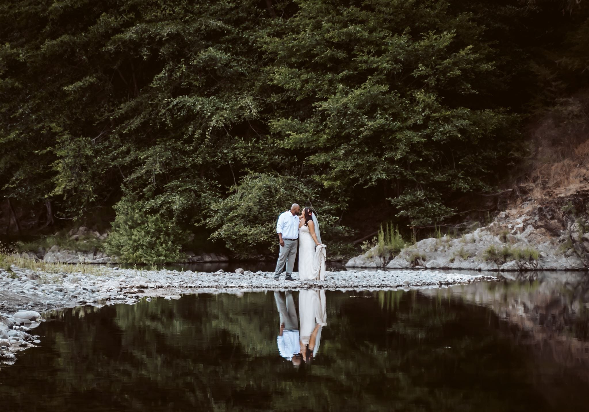 A couple standing by a calm river with dense forest in the background.
