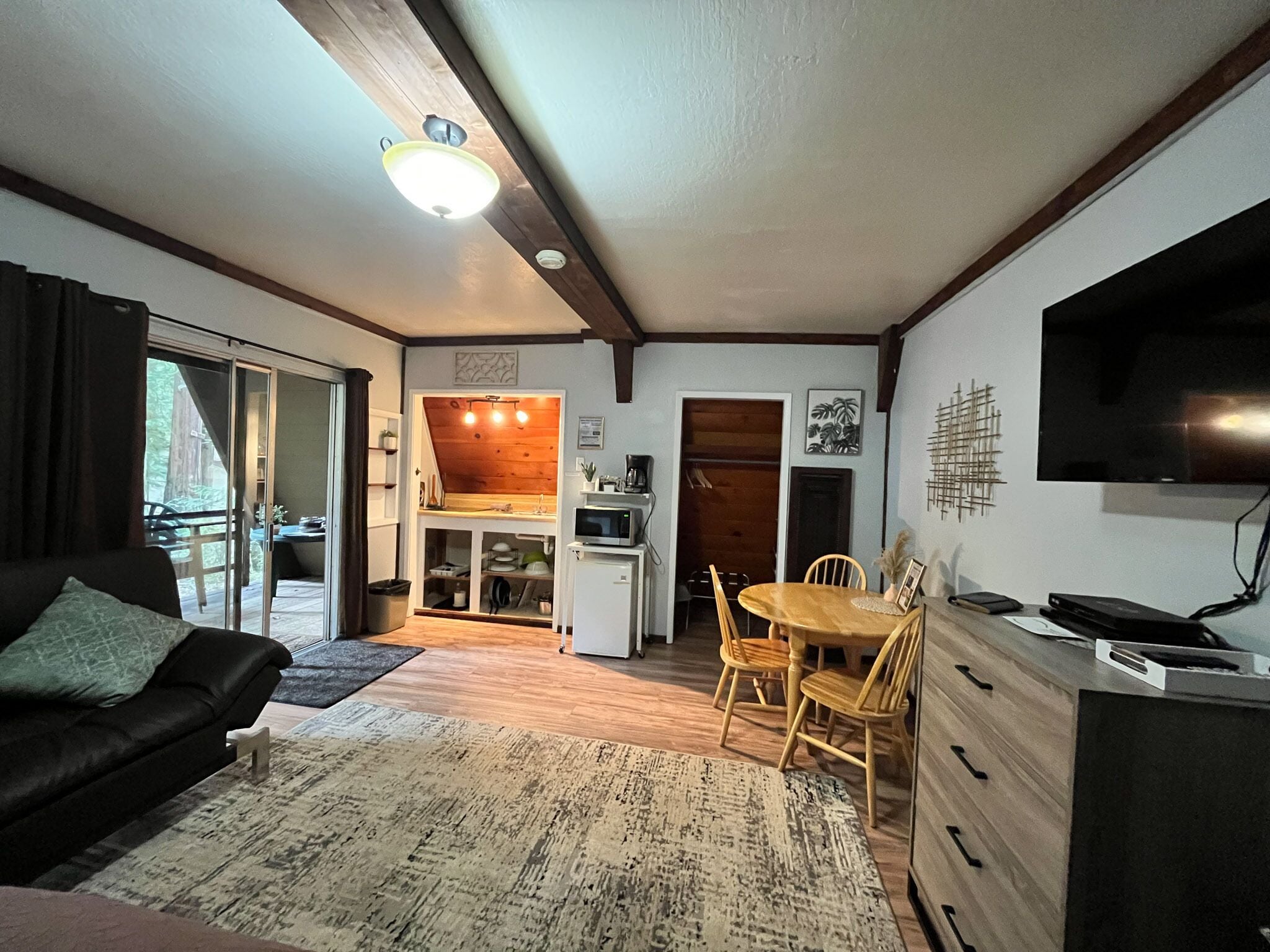 A cozy living room with a sofa, a dining table with chairs, a kitchen area with a microwave, and a TV mounted on the wall.
