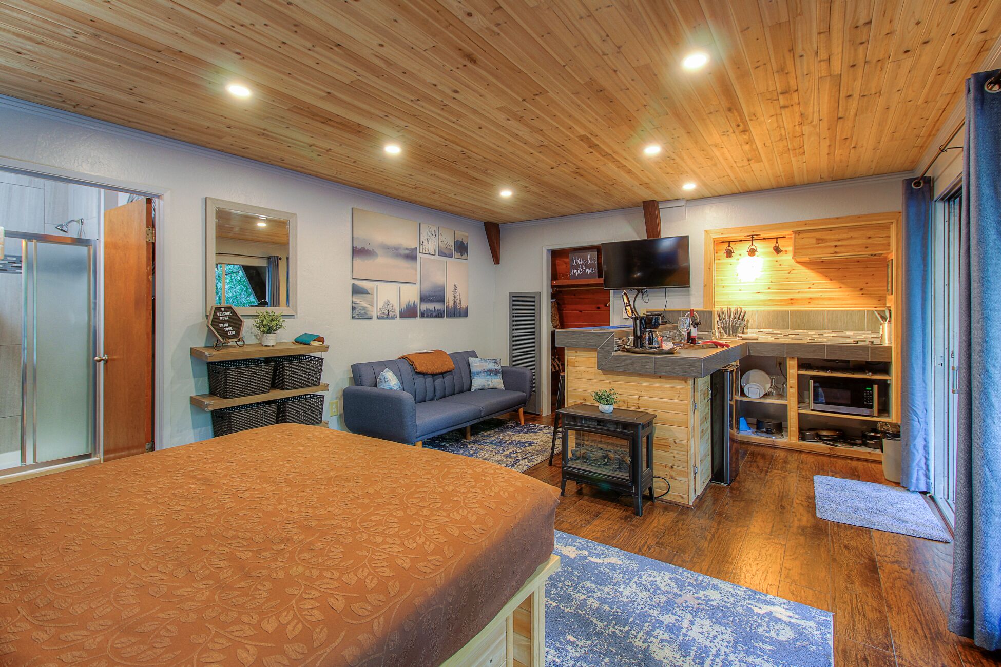 A cozy living room with a sofa, a coffee table, and a television. There is a wooden ceiling and a kitchen area with a microwave and a sink.