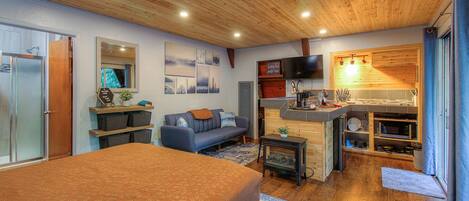 A cozy living room with a sofa, a coffee table, and a television. There is a wooden ceiling and a kitchen area with a microwave and a sink.