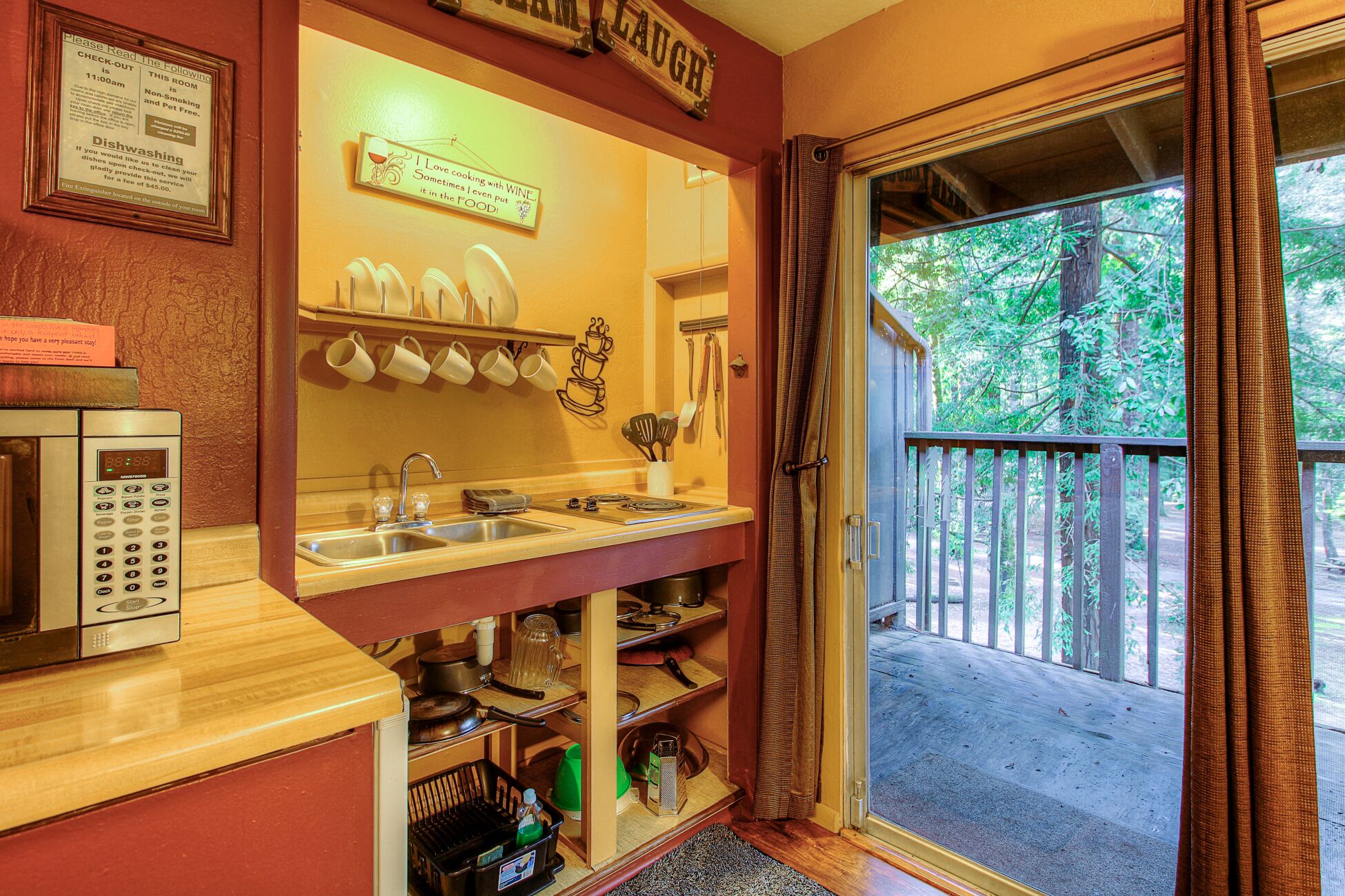 A compact kitchenette with a sink, stove, and microwave. There are mugs and utensils hanging above the sink, and a sign above the door.