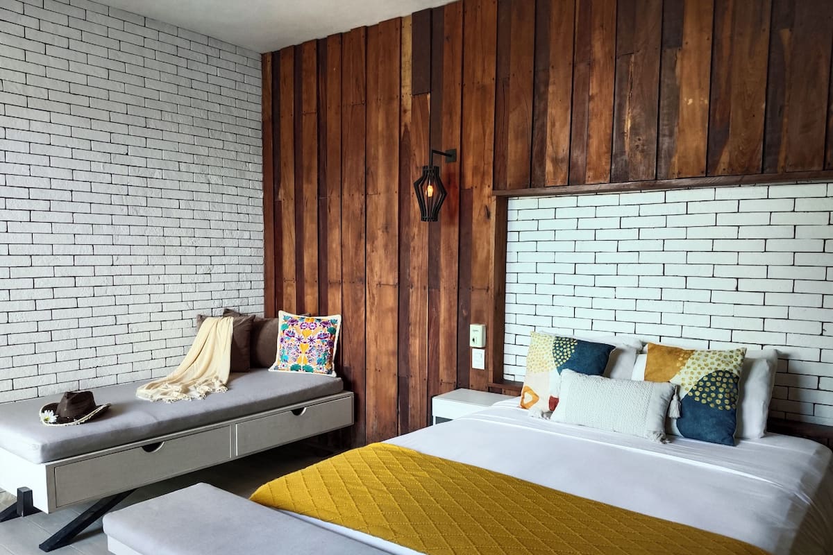 Quarto moderno com parede em padrão tijolo, painéis de madeira, cama com várias almofadas, um banco com manta e mesa de cabeceira com abajur.