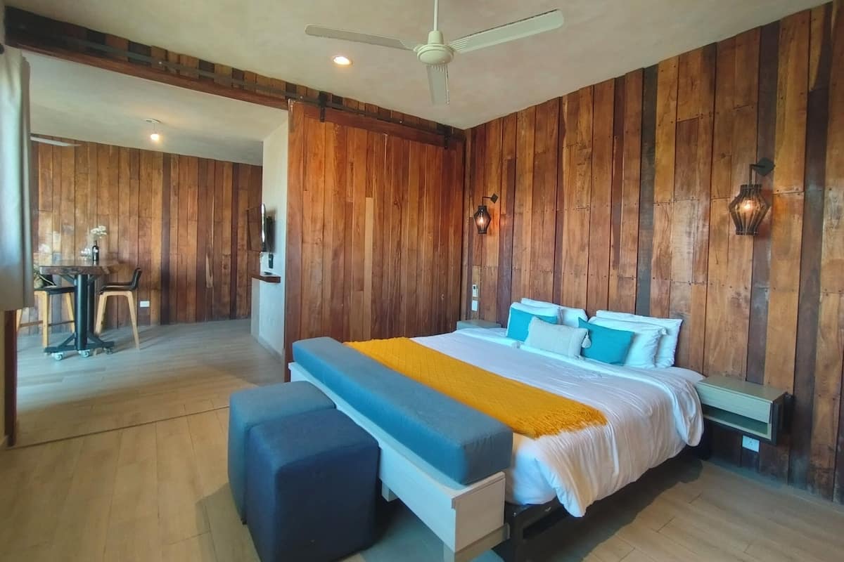 Quarto com cama, mesas de cabeceira, escrivaninha e cadeira, e ventoinha no teto.