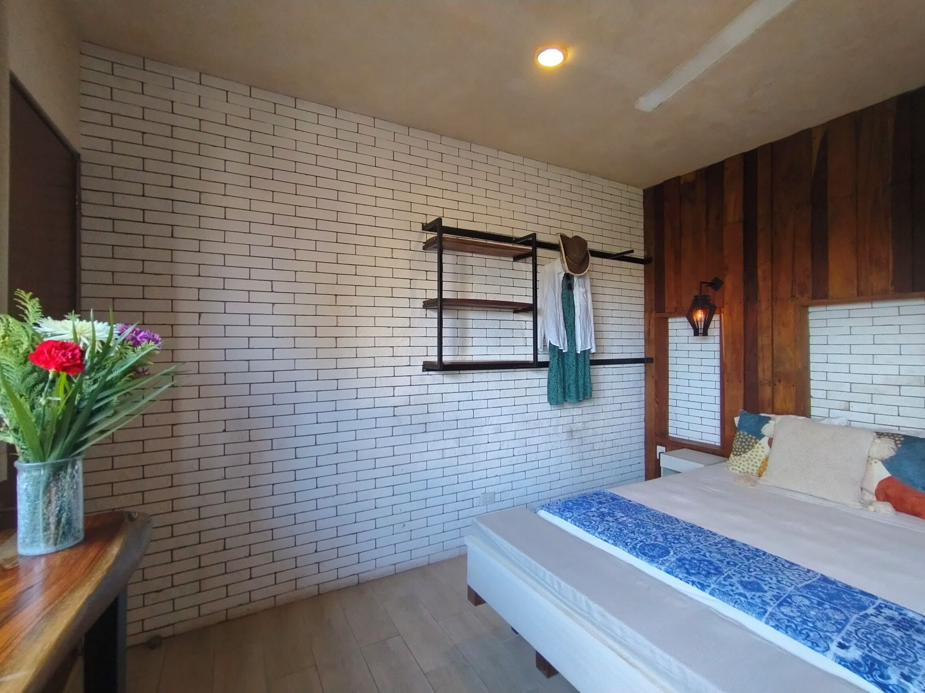 Un dormitorio con pared de ladrillo, una cama con colcha azul y blanca estampada, cabecera de madera, un perchero y un jarrón con flores sobre una mesa de madera.
