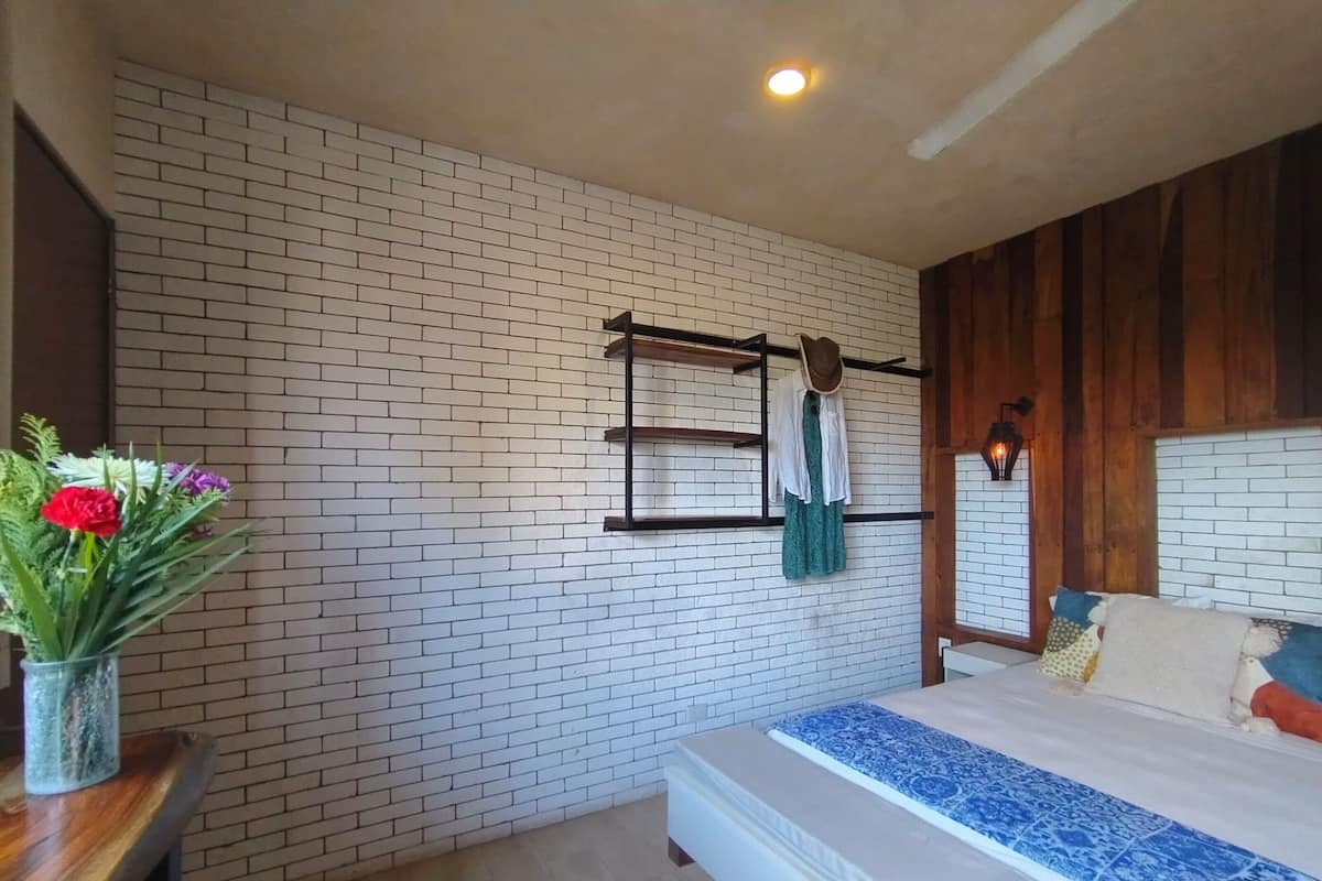 Quarto com parede de tijolos, cama com colcha azul e branca estampada, cabeceira de madeira, cabideiro suspenso e um vaso com flores sobre uma mesa de madeira.