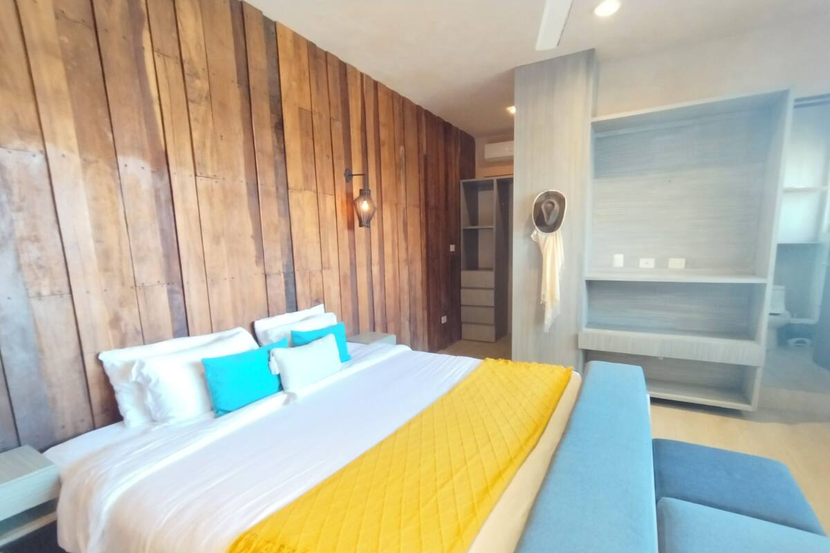 Quarto de hotel moderno com parede de madeira, cama com roupa de cama branca e azul, um cobertor amarelo e um sofá azul.