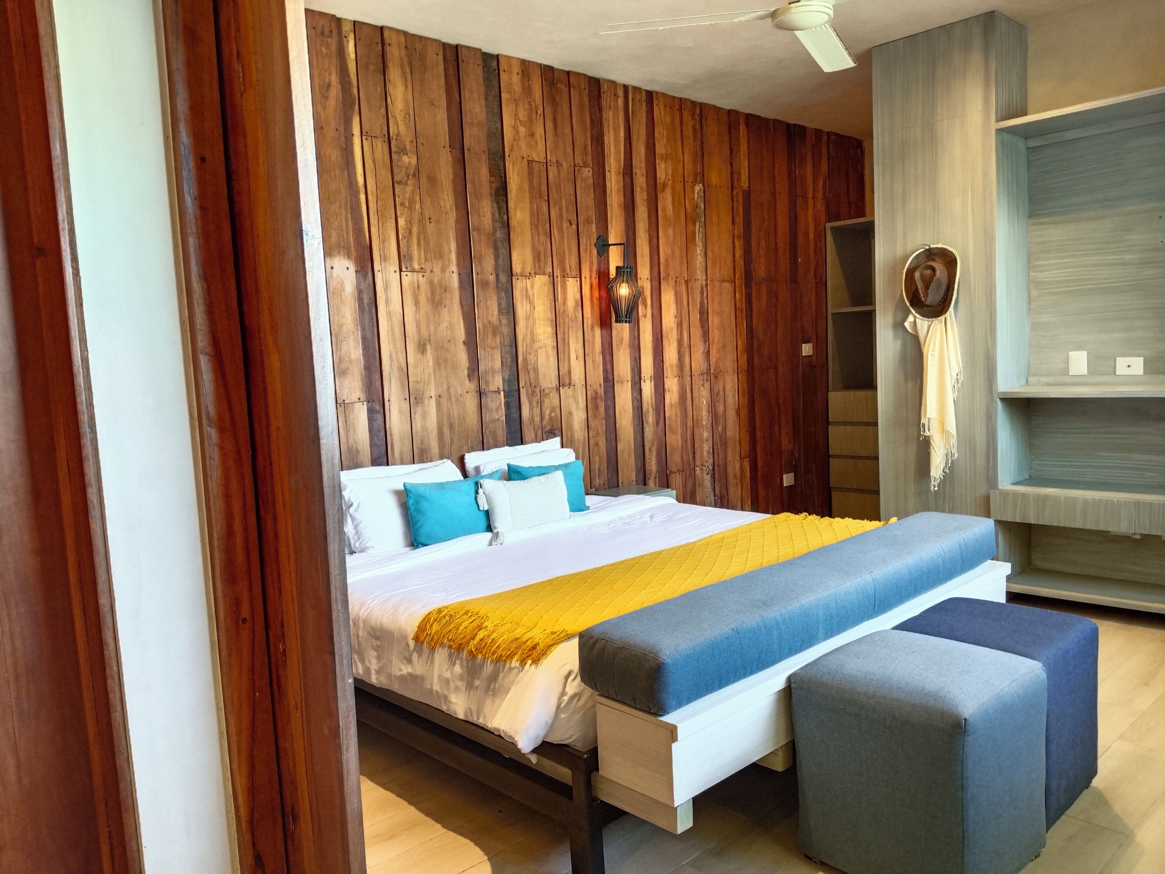 Quarto moderno com parede de painéis de madeira, cama com roupa de cama branca e azul, manta amarela e um banquinho cinza.
