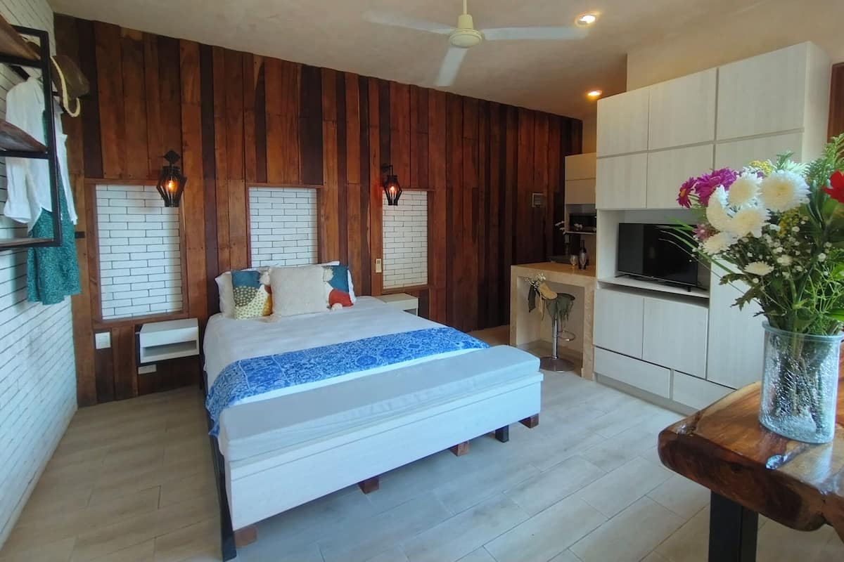 Quarto com cama, paredes de madeira, ventilador de teto, uma pequena mesa com um vaso de flores e uma área de cozinha compacta.
