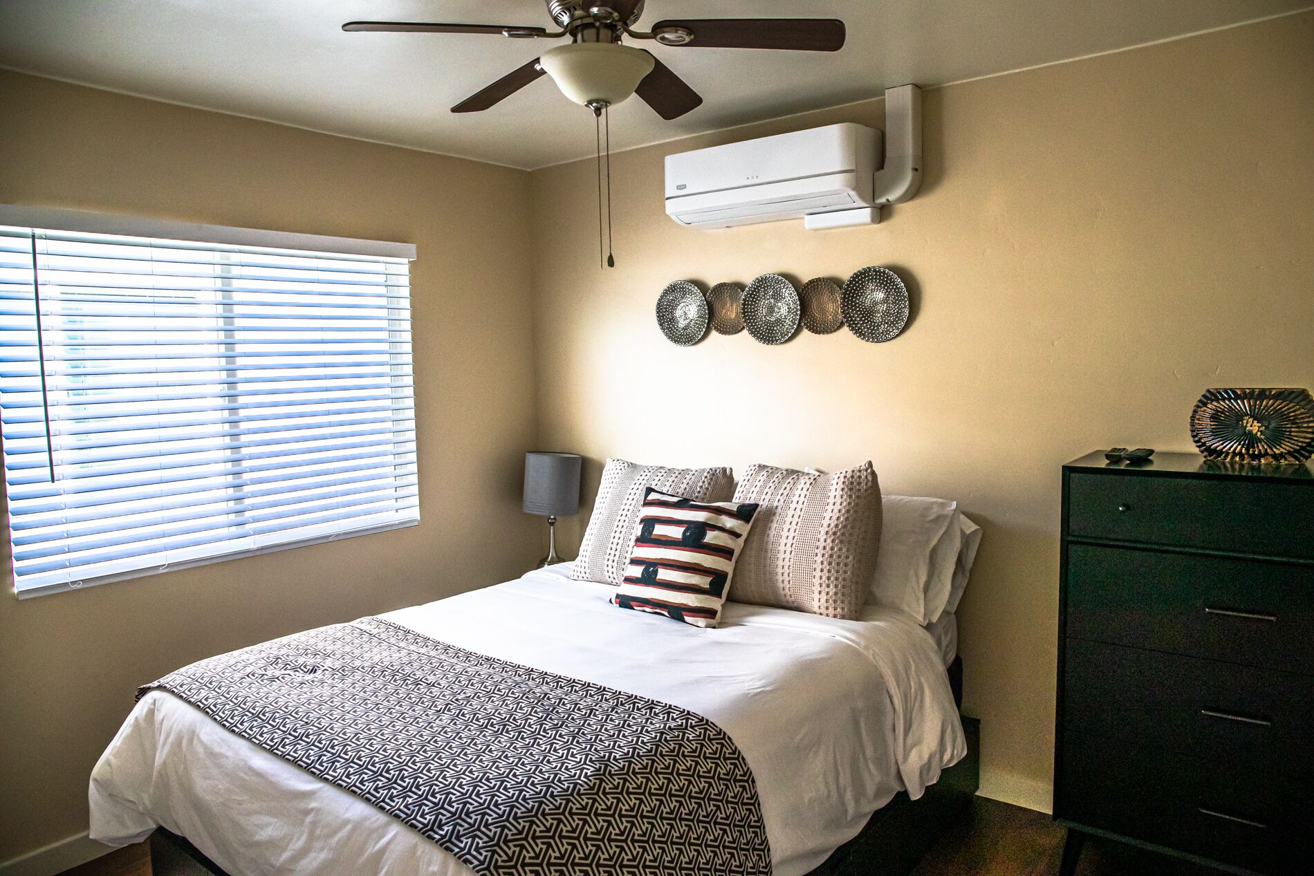 A bedroom with a bed, a ceiling fan, an air conditioner, a window with blinds, a wall-mounted decorative plate, and a green dresser.