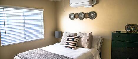 A bedroom with a bed, a ceiling fan, an air conditioner, a window with blinds, a wall-mounted decorative plate, and a green dresser.