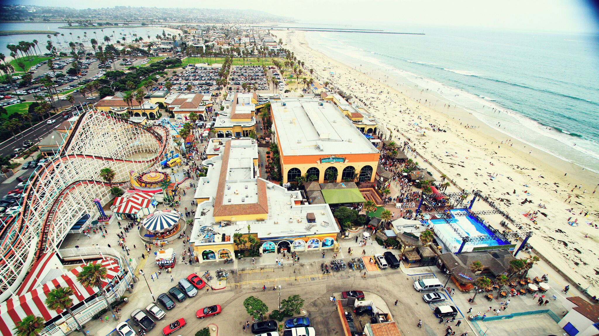 A coastal cityscape with a beach, a roller coaster, and a large building with arches.