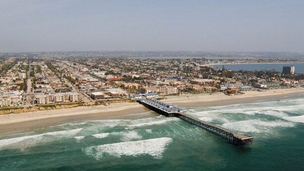 Vista aérea de una ciudad costera con un muelle largo que se extiende hacia el océano, rodeada de edificios residenciales y una playa.