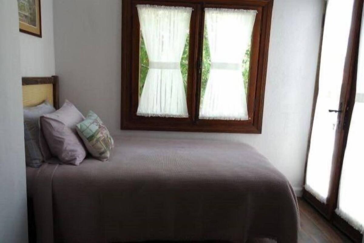 A single bed with a brown bedspread and two pillows, a window with white curtains, and a wooden frame.