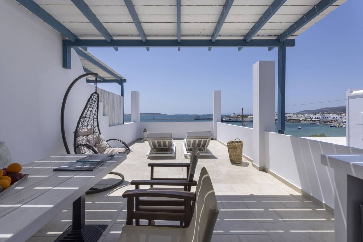 A balcony with a view of the sea, a table with fruits, two chairs, and a hanging chair.