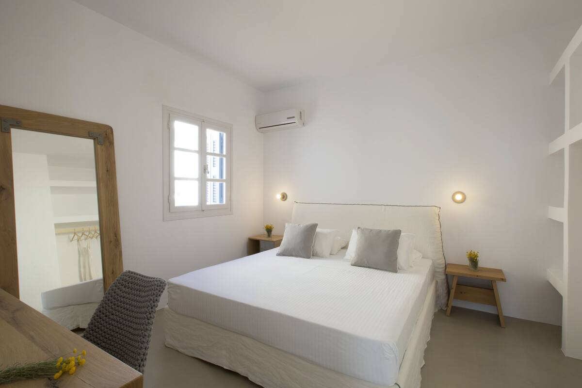 A bedroom with a large bed, a wooden nightstand with a lamp, a small table with flowers, and a mirror on the wall.