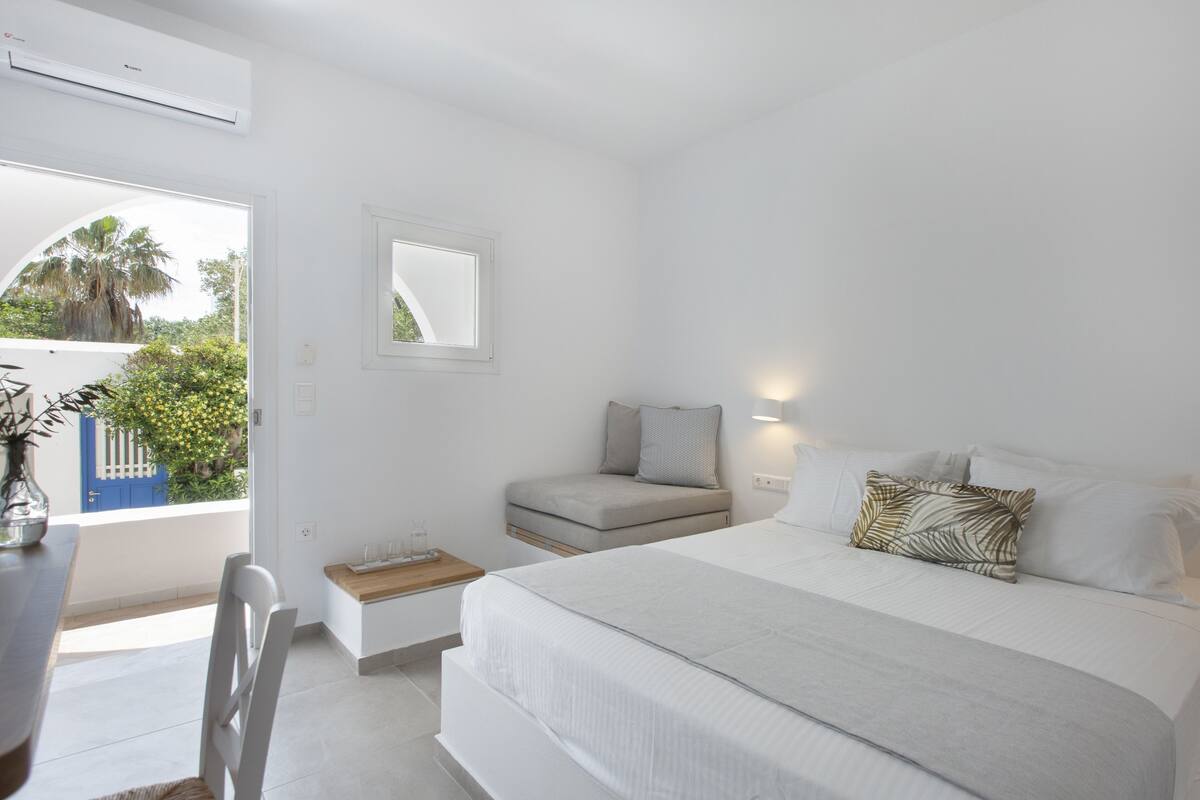 A modern bedroom with a large bed, a small seating area, a window with a view of greenery, and a white color scheme.