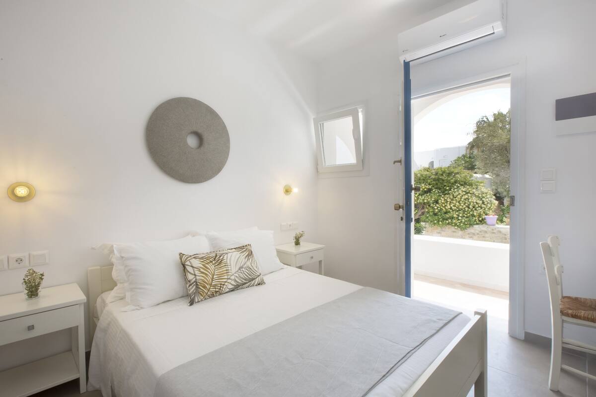 A bedroom with a bed, bedside tables, a wall-mounted light fixture, a window, and a view of the outdoors.