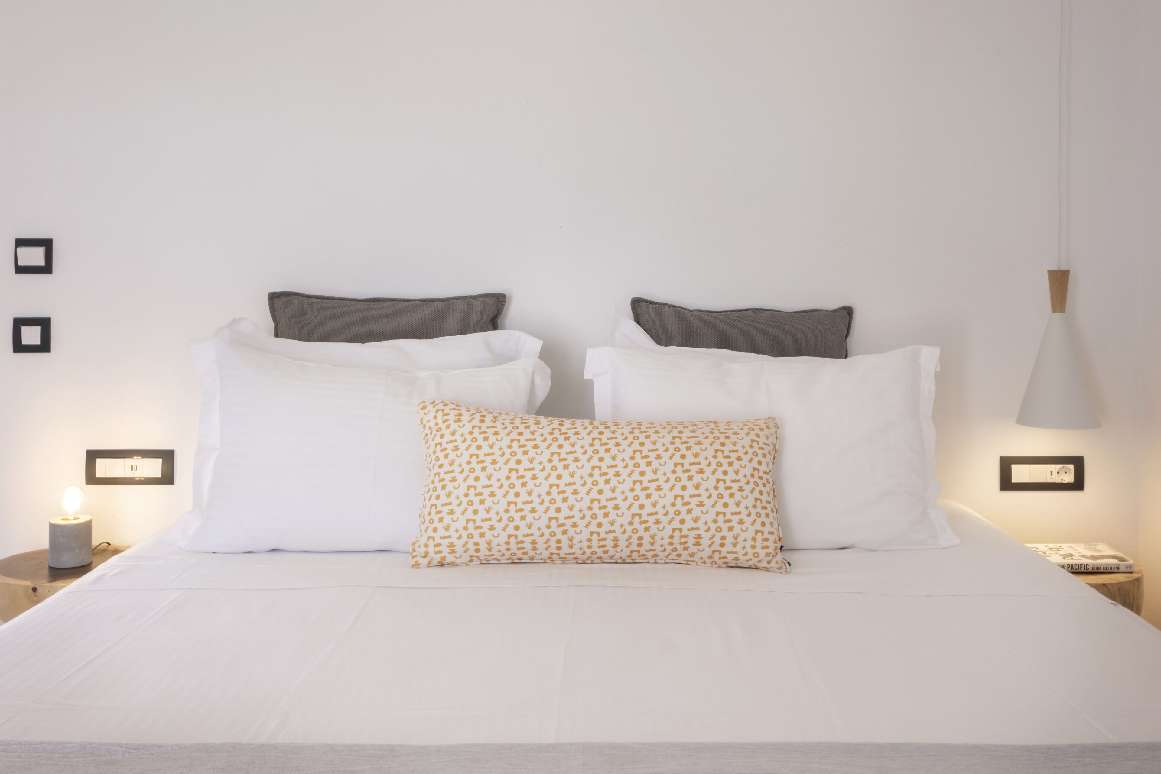 A neatly made bed with white and patterned pillows, a bedside lamp, and a small table with a book.