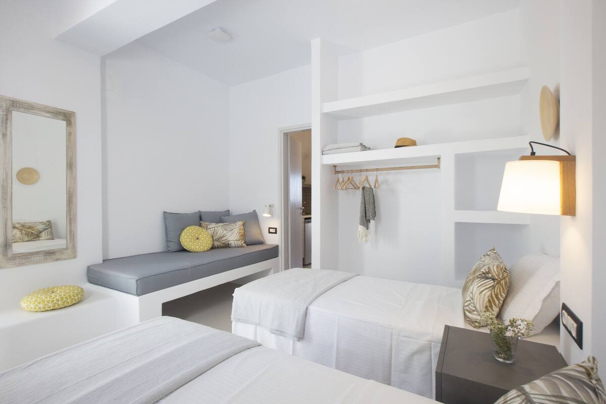 A modern hotel room with a built-in bench, two beds, a bedside table with a lamp, and a wardrobe with hanging clothes.