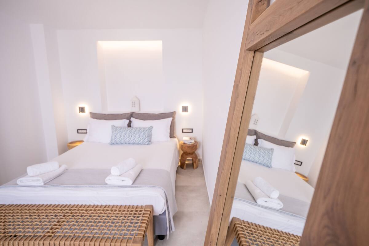 A hotel room with two beds, a wooden bench, a small stool, and a mirror.