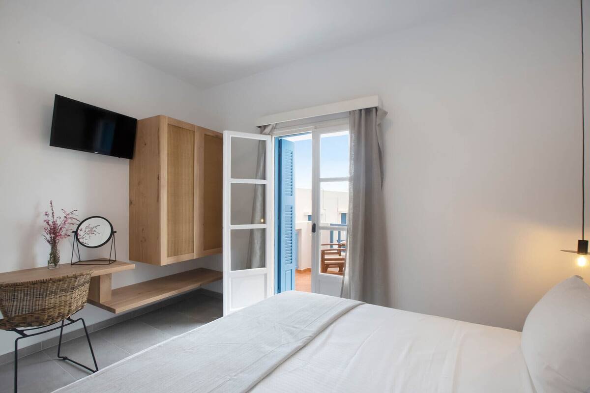 A bedroom with a bed, a TV mounted on the wall, a wooden cabinet, a wicker chair, and a view of the outside through an open door.