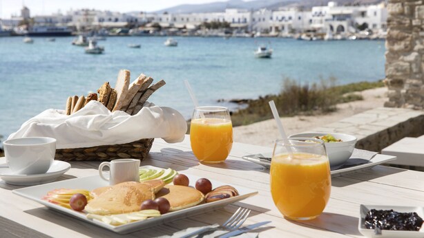 A breakfast table with croissants, fruit, pancakes, and orange juice, overlooking a harbor with boats and white buildings. - Kouros Village (Antiparos)