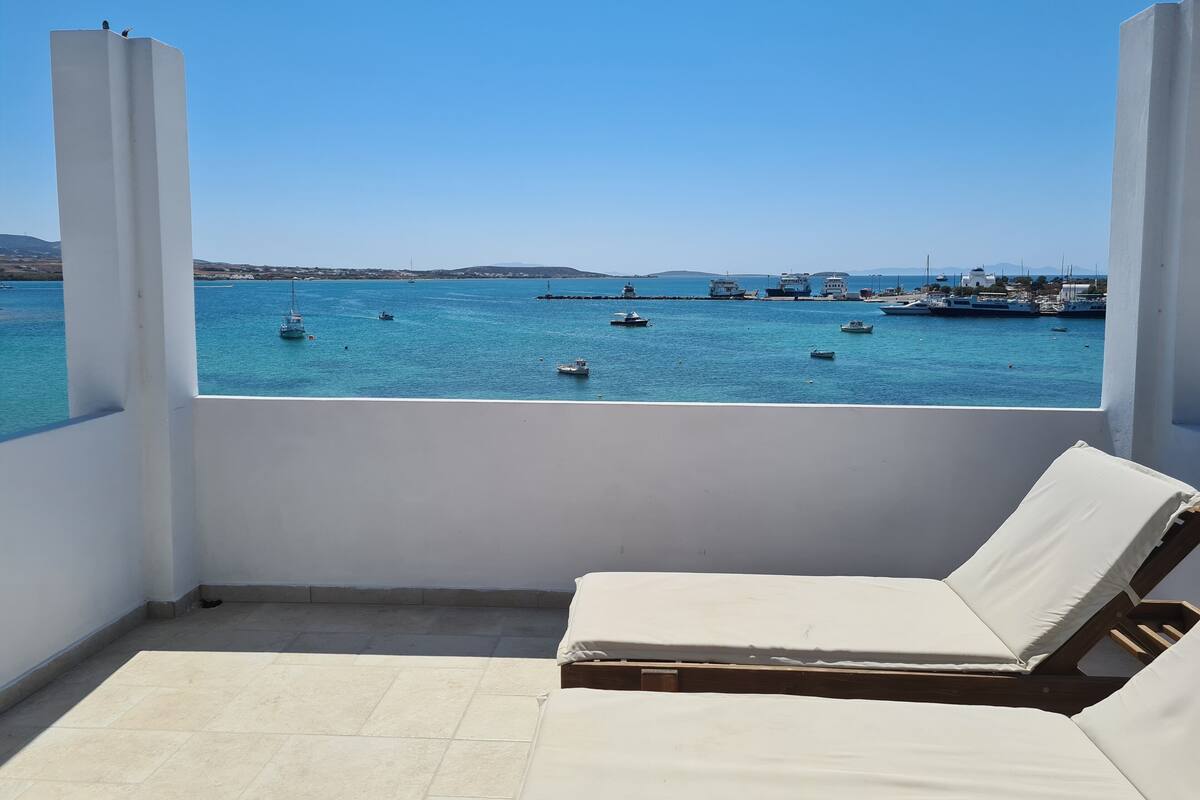 A balcony with a white cushioned lounge chair, a tiled floor, and a view of the sea with boats and a lighthouse in the distance.