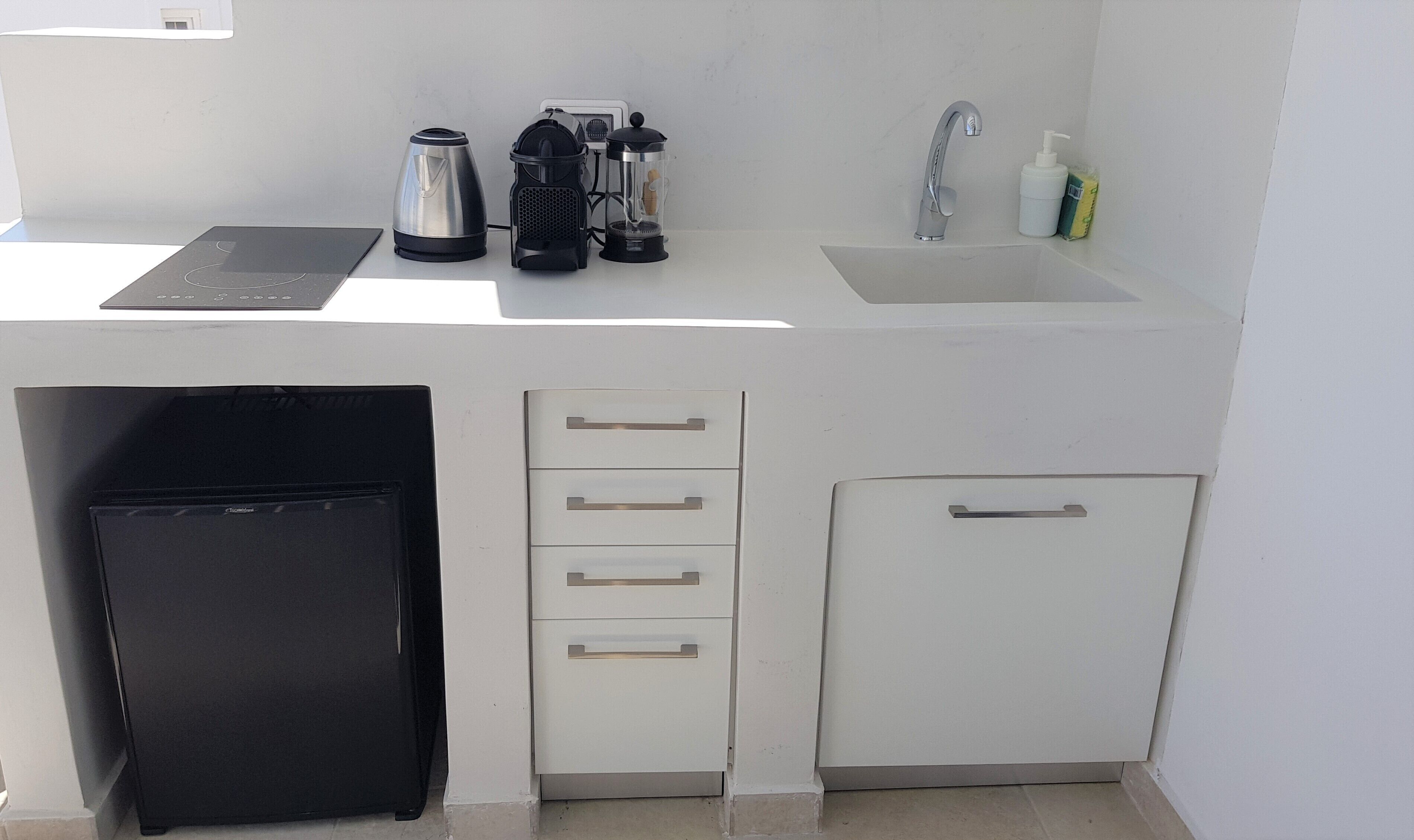 Una kitchenette compacta con un refrigerador negro, una cafetera, un molinillo, una tetera y un lavamanos con grifo.