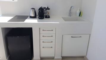 Una kitchenette compacta con un refrigerador negro, una cafetera, un molinillo, una tetera y un lavamanos con grifo.
