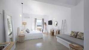 A modern, minimalist bedroom with a large bed, a sofa, a TV, and a view of the outdoors. - Kouros Village (Antiparos)