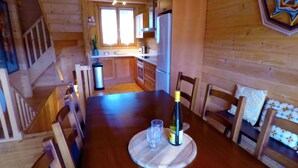 Interior de madera con una mesa de comedor preparada para dos personas, una cocina al fondo y una escalera que sube a otro nivel.