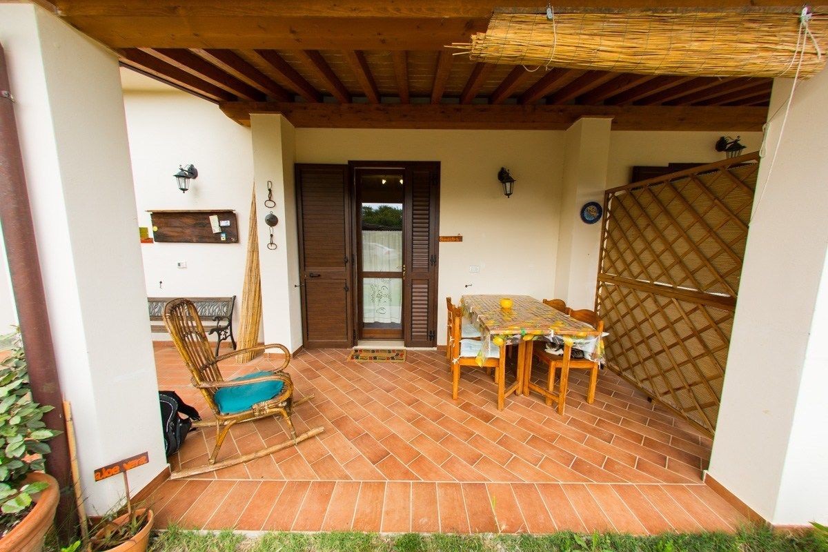 Un patio techado con suelo de baldosas, una mesa de madera con sillas, una silla de mimbre y una puerta con ventanal.