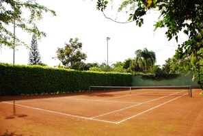 An outdoor tennis court surrounded by greenery and a hedge.
