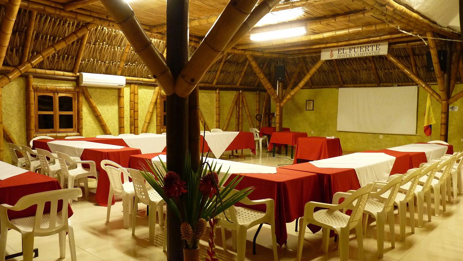 Un salón de banquetes con mesas dispuestas para comer, sillas de plástico blancas y una estructura de techo de bambú.