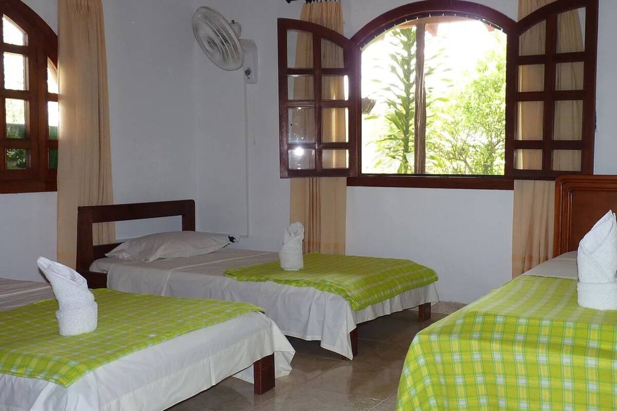 A hotel room with three single beds, each with a green checkered bedspread and a white towel on the pillows. There is a window with wooden shutters and a fan mounted on the wall.