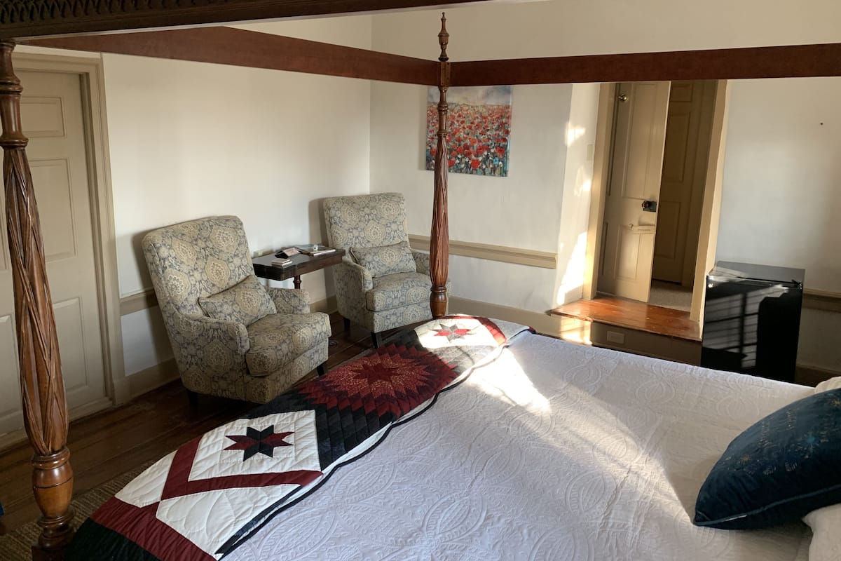 A bedroom with a four-poster bed, two armchairs, a small table, and a painting on the wall.