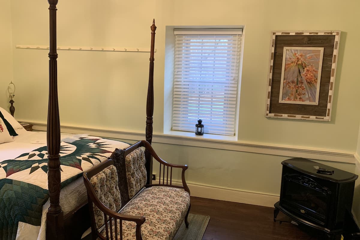 A bedroom with a four-poster bed, a floral upholstered chair, a small window with blinds, a framed picture on the wall, and a black dresser.