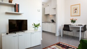 Une cuisine moderne avec des meubles blancs, une table à manger avec des chaises, un téléviseur à écran plat posé sur un meuble et un tableau encadré au mur.