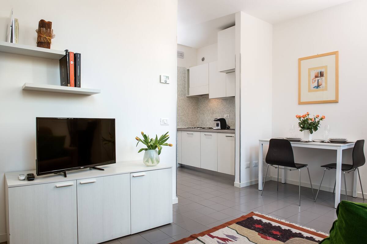 A modern kitchen with white cabinets, a dining table with chairs, a flat-screen TV on a cabinet, and a framed picture on the wall.