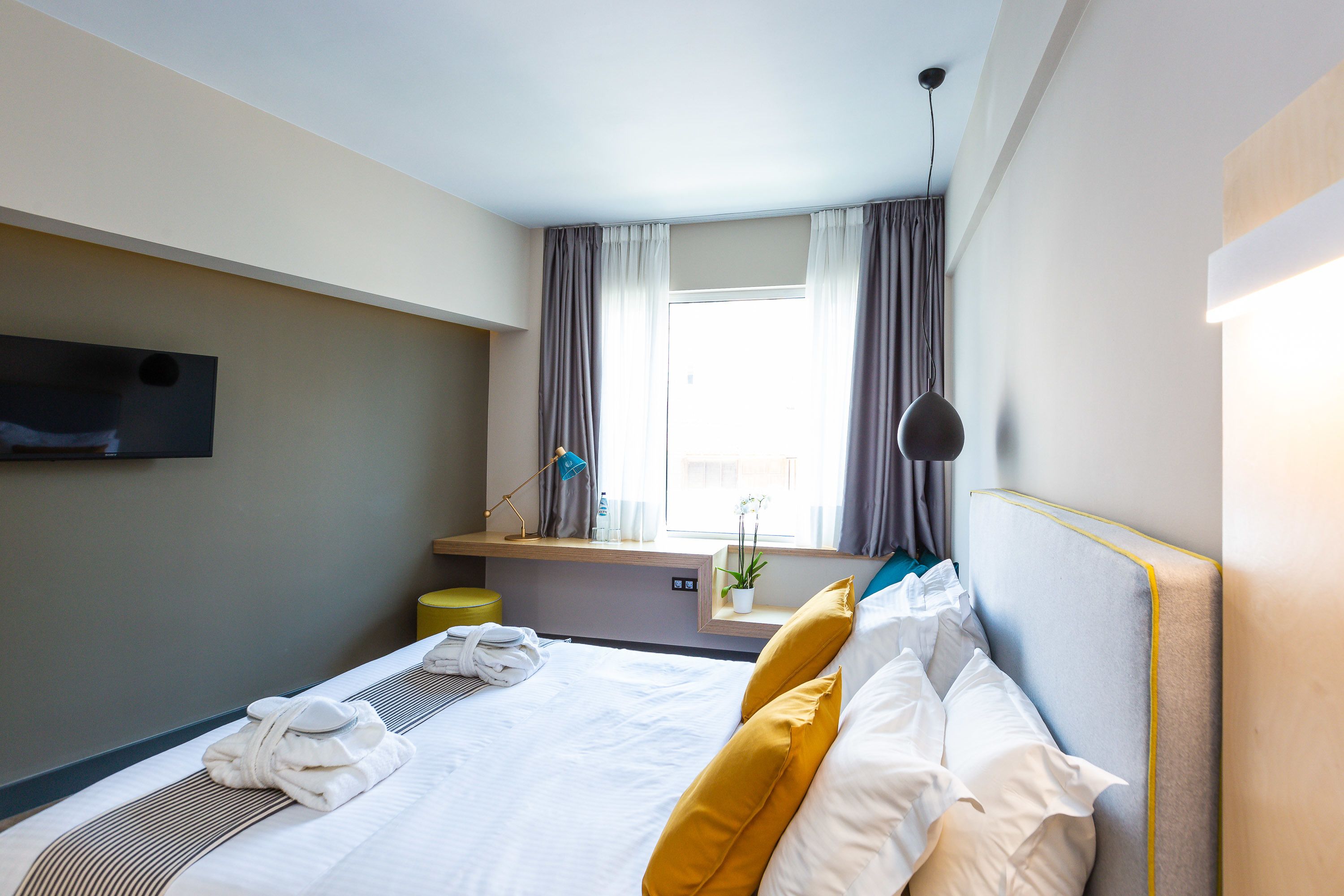 Een hotelkamer met een bed, een aan de muur bevestigde televisie, een raam met gordijnen en een nachtkastje met een lamp en een plant.