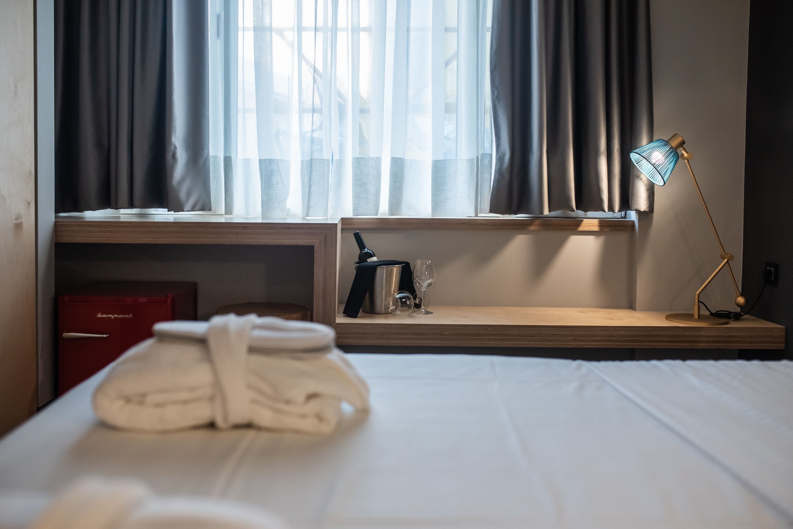 Une chambre d’hôtel avec un lit bien fait, une table de chevet avec une lampe, une boîte rouge et une fenêtre avec des rideaux.