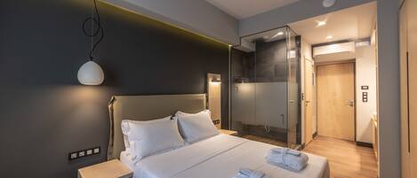A modern hotel room with a large bed, bedside tables, a glass shower enclosure, and a visible bathroom door.