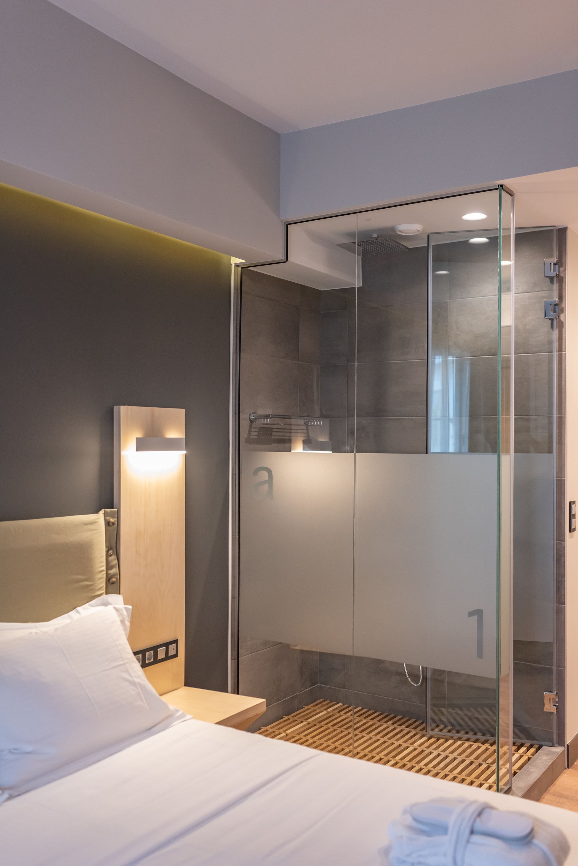 A modern hotel room with a glass-enclosed shower, a neatly made bed, and a wall-mounted light fixture.