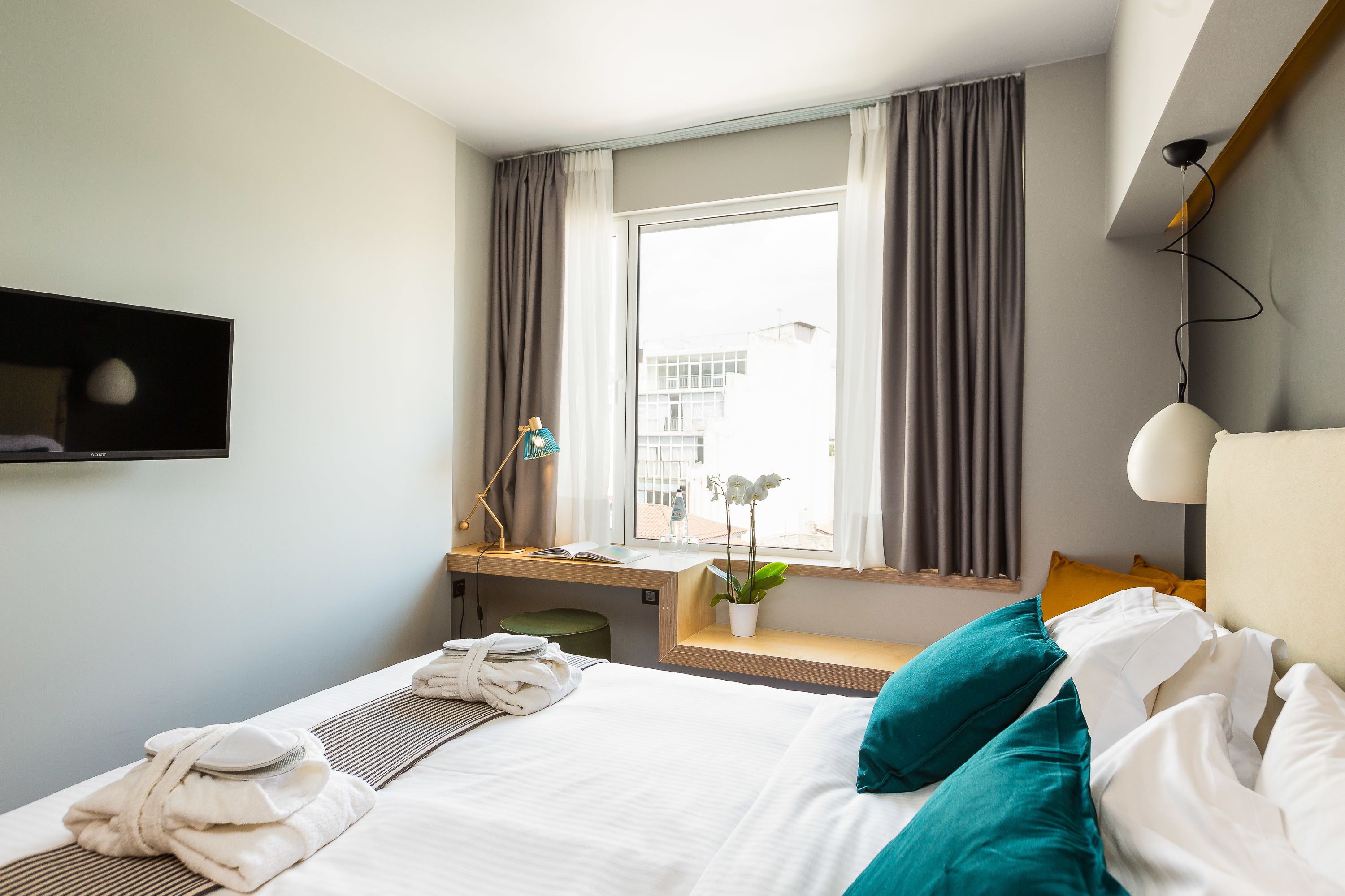 Ett hotellrum med en säng, ett skrivbord, ett fönster med gardiner, en krukväxt och en tv som är monterad på väggen.