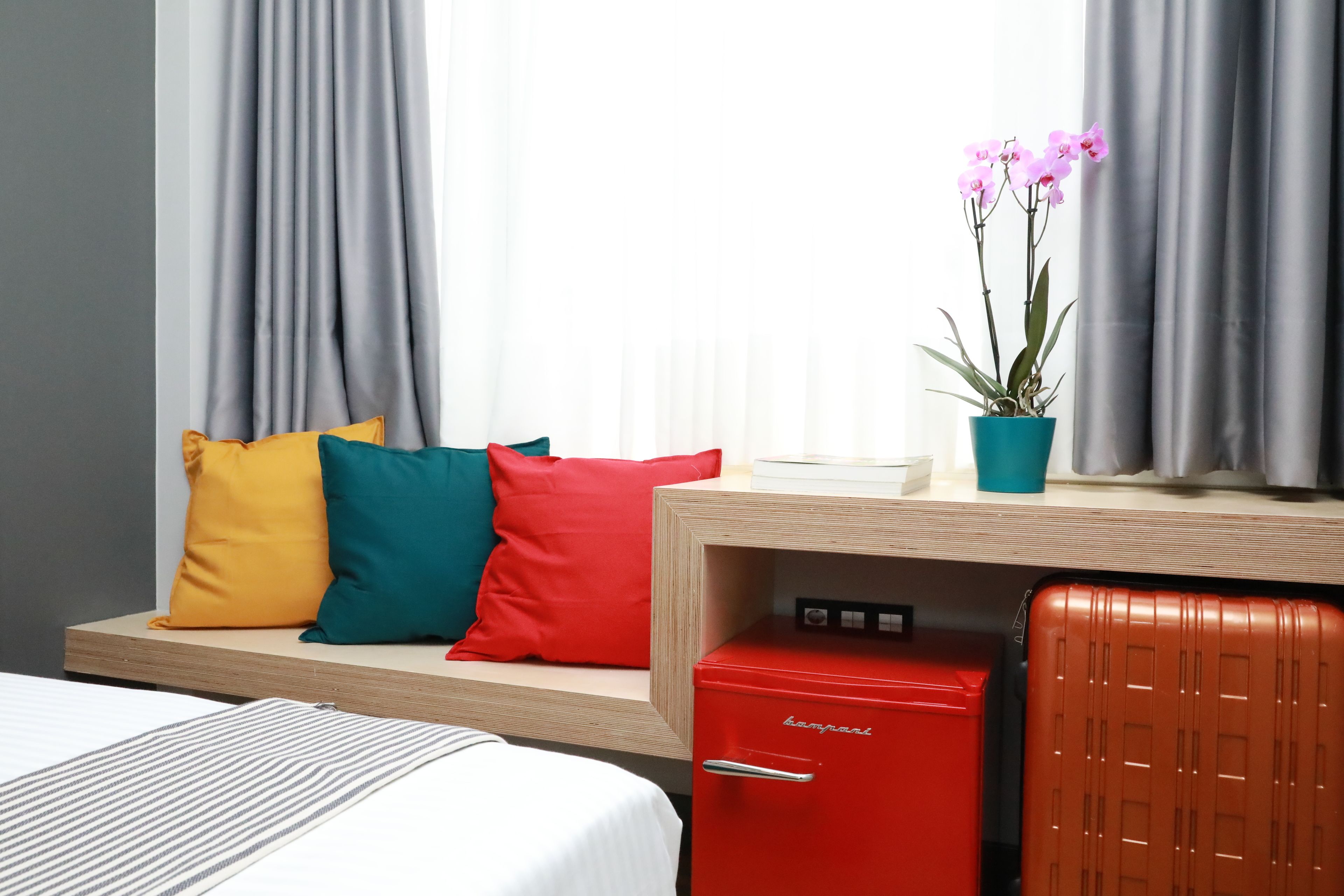 A hotel room with a bed, a desk with a red filing cabinet, a potted orchid, and a window with grey curtains.
