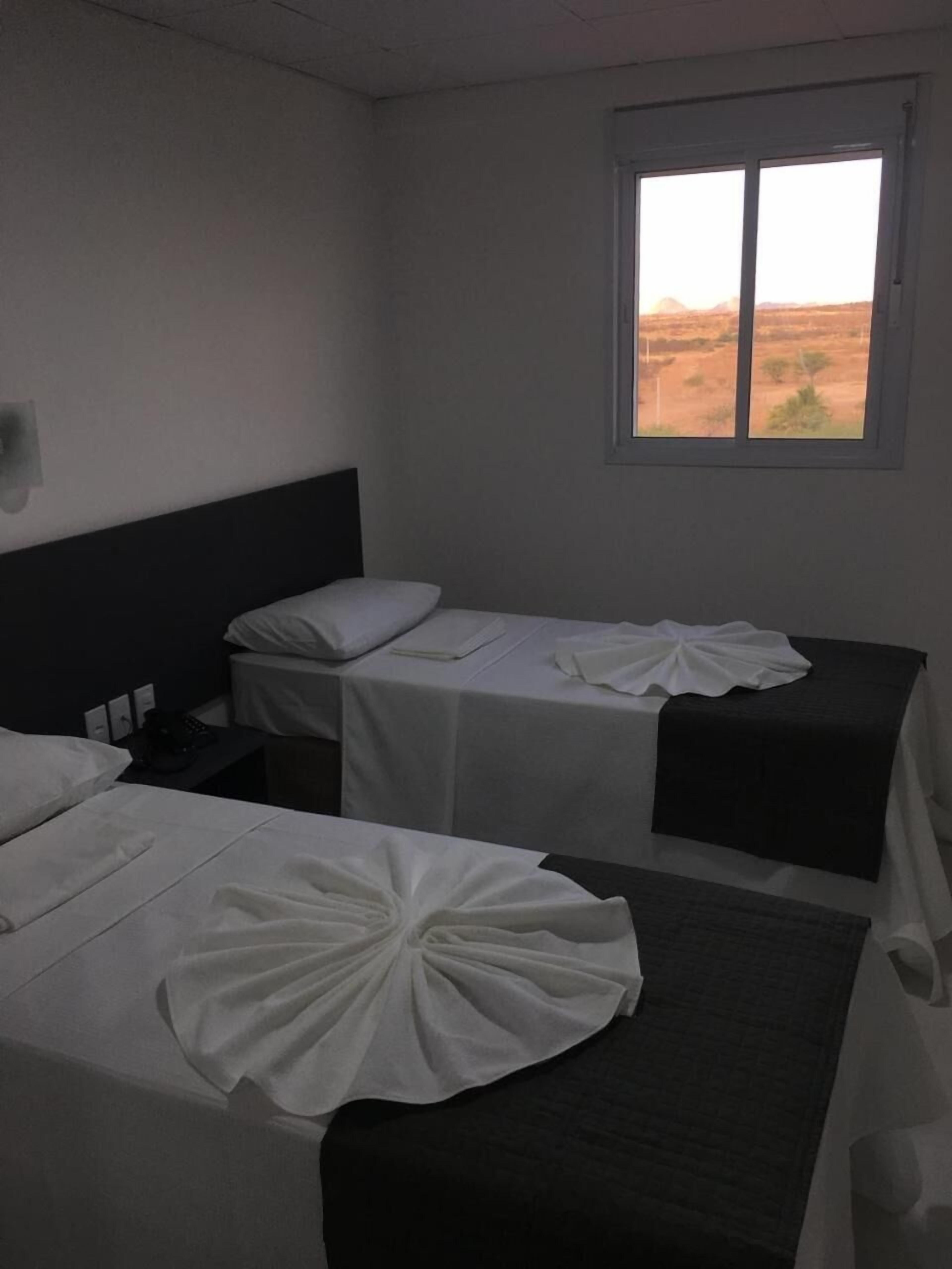 A room with two single beds, a small table, a nightstand, and a window with a view of a desert landscape.