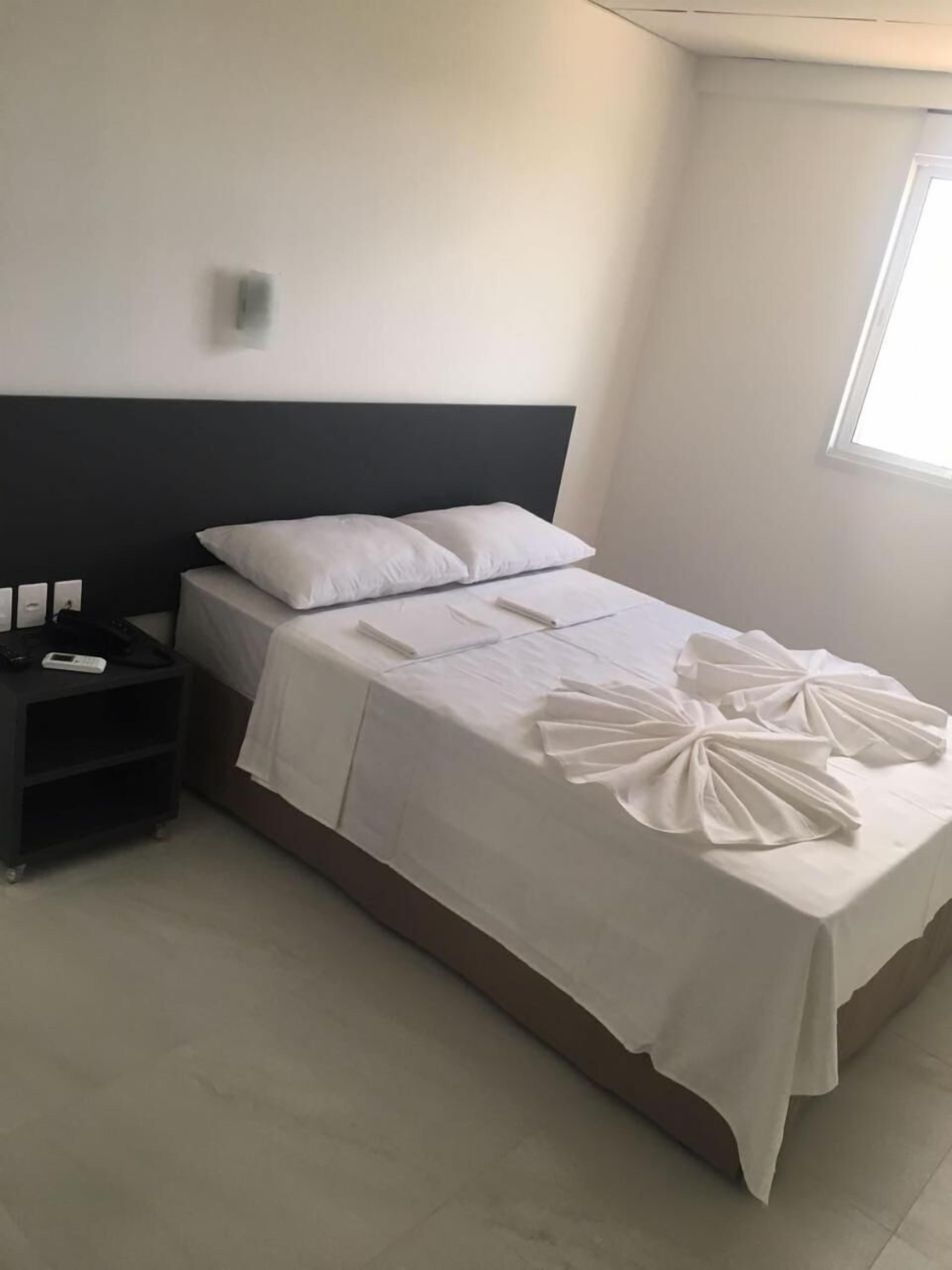 Cama bem arrumada com lençóis brancos e almofadas decorativas, mesa de cabeceira com telefone e carregador, e janela que proporciona luz natural.