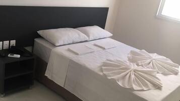 Cama bem arrumada com lençóis brancos e almofadas decorativas, mesa de cabeceira com telefone e carregador, e janela que proporciona luz natural.
