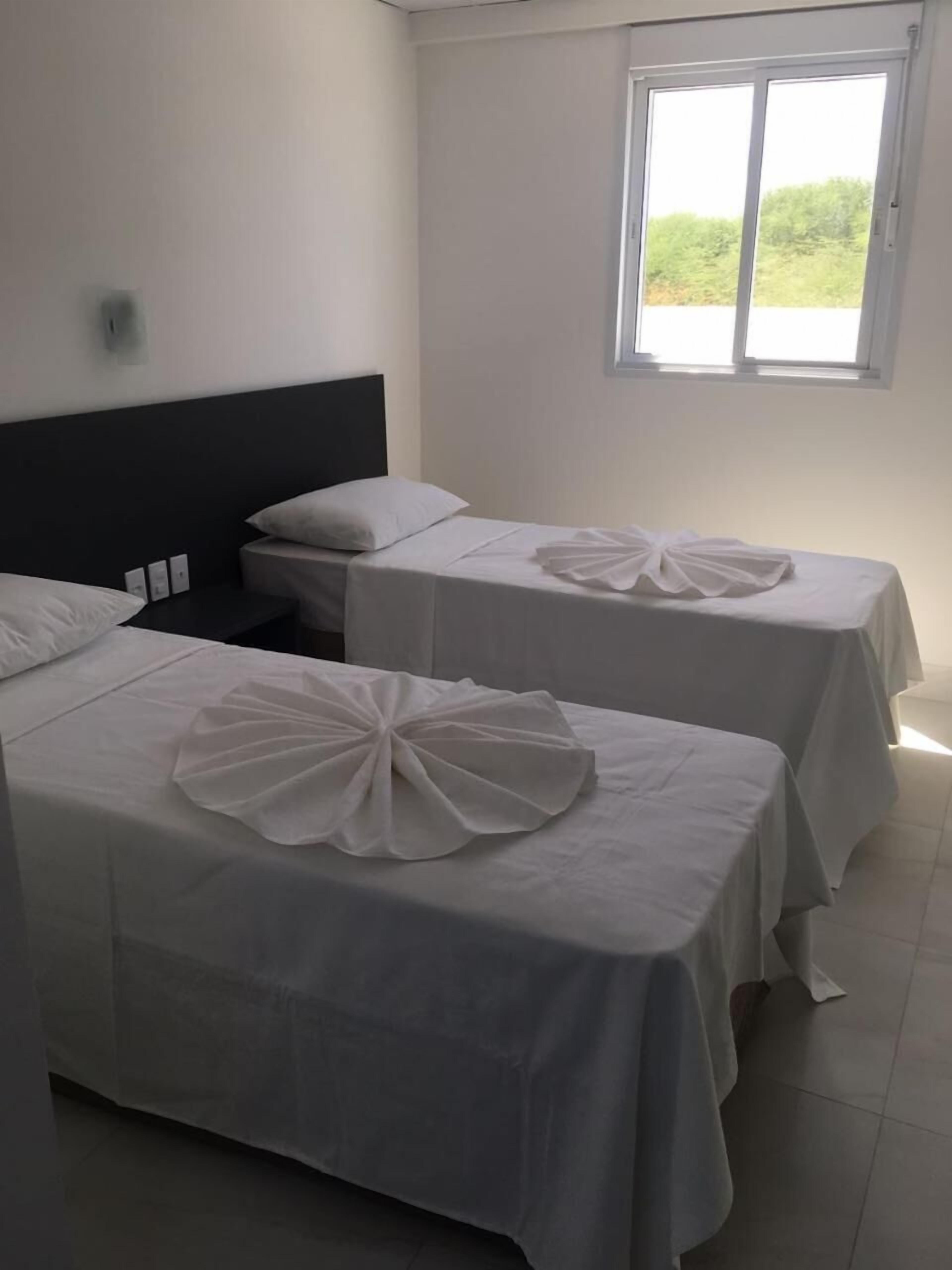 A hotel room with two single beds, each with a white bedspread and a decorative white bow on the bed. There is a window with a view of greenery outside.
